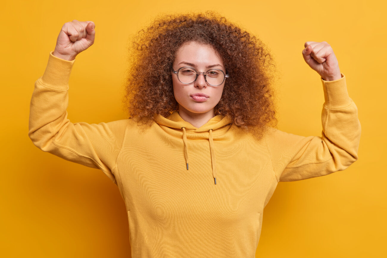 Serious beautiful European with curly hair looks confident at camera raises arms shows muscles wears spectacles and sweatshirt isolated over yellow background gets strong brags about strength